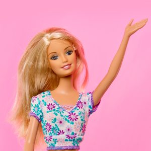 Doll raises arm, wearing a floral top with blonde hair, set against a pink background.