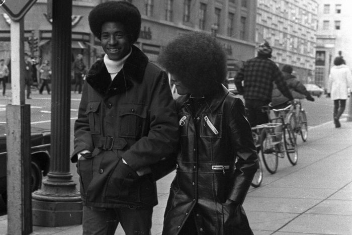 Two people walk on a city sidewalk, wearing stylish 1970s outfits, with a street sign and bicycles in the background.