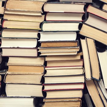 Books stacked horizontally, forming uneven piles, with varied colors and sizes, creating a chaotic, cluttered appearance. No visible text or titles.