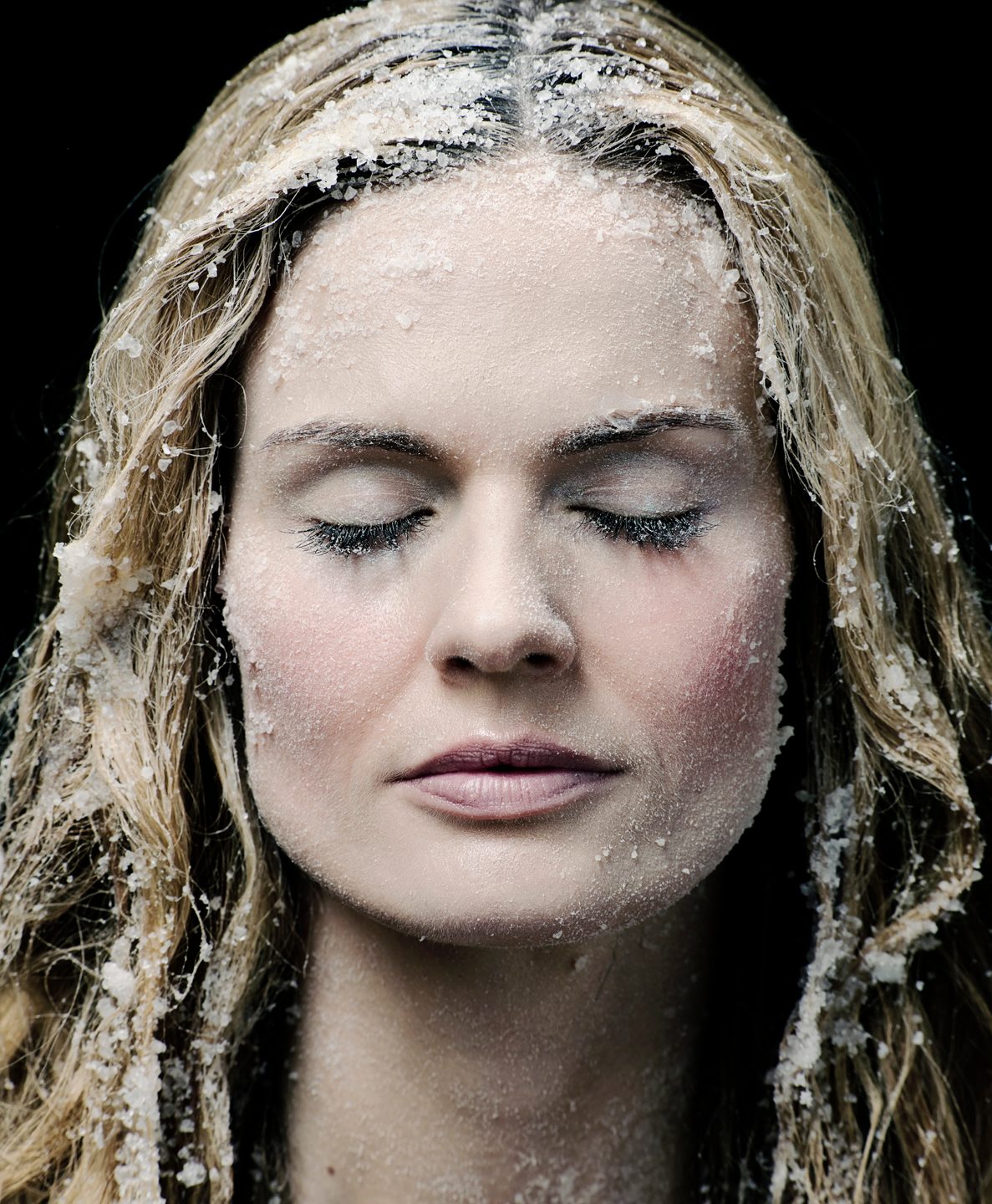 A woman with closed eyes appears peaceful, snow covering her hair and face, against a dark background.