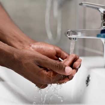Hands catch water running from a faucet in a white sink against a blurry bathroom background.