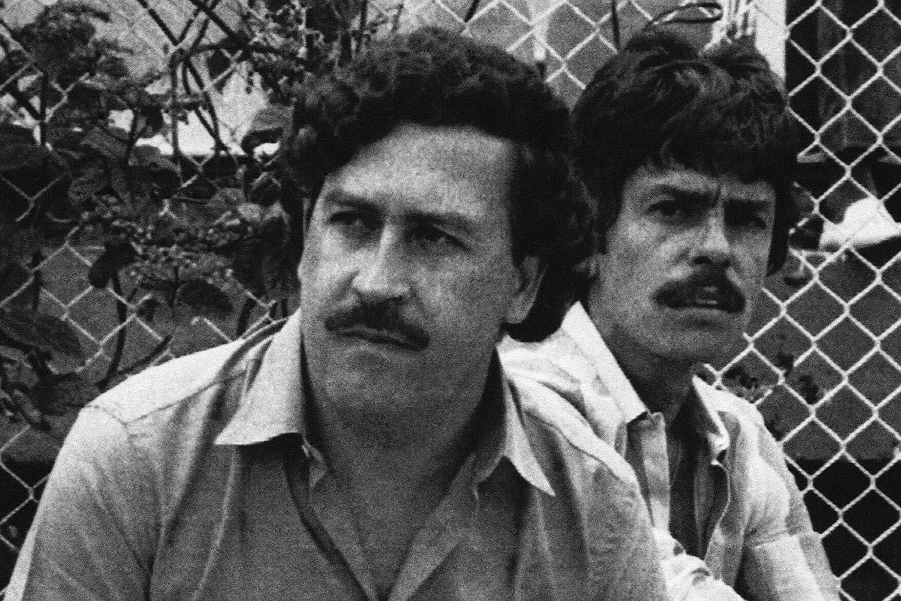 Two men sit beside a chain-link fence, one holding a cigarette, appearing contemplative in an outdoor setting.