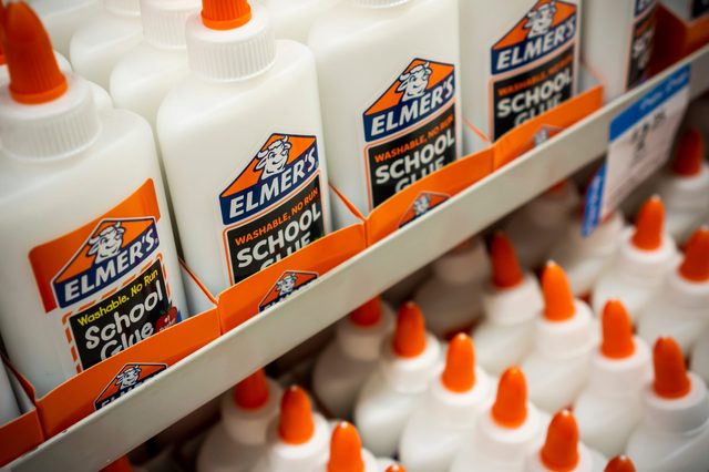 Glue bottles sit on store shelf, arranged with orange caps visible; labeled "Elmer's School Glue" with "Washable, No Run" text.