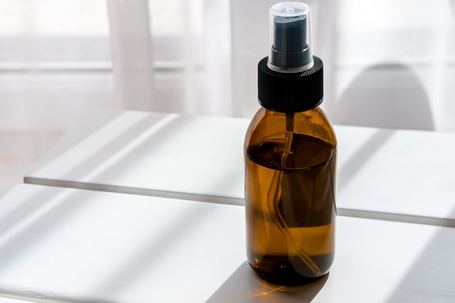 A brown spray bottle stands upright on a white surface, with soft light streaming through sheer curtains in the background.