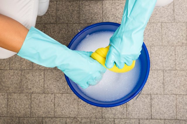 Gloved hands wring a yellow sponge over a blue bucket filled with soapy water, on a tiled floor.