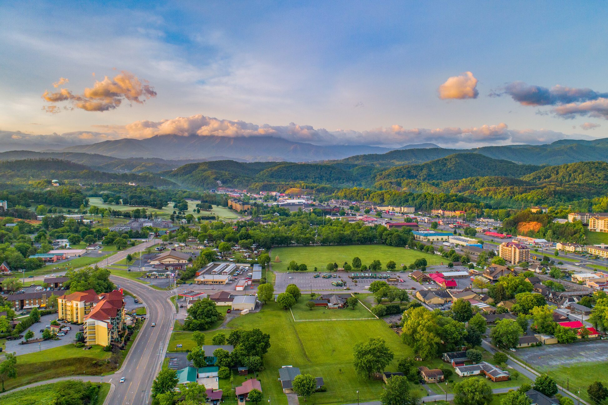 Aerial view shows a small town amidst lush greenery and distant mountains under a clear sky with scattered clouds.