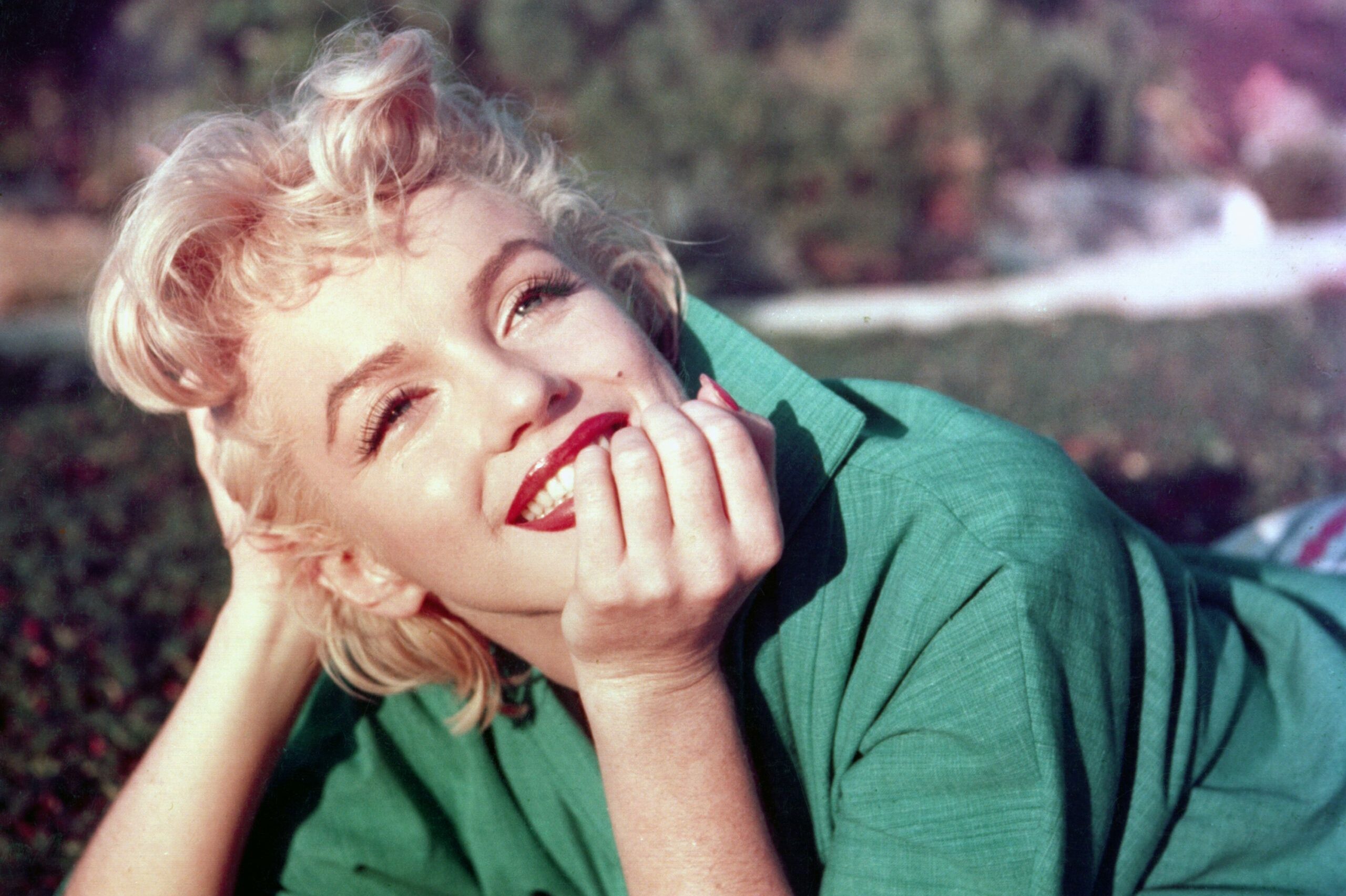 Person smiling and reclining on grass, wearing a green shirt, with a hand touching their face. Background shows blurred foliage.