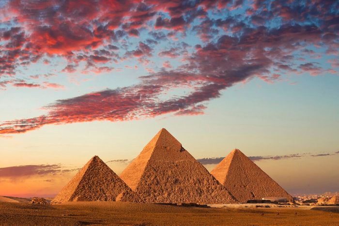Pyramids stand still under a vibrant, colorful sunset sky, surrounded by desert terrain and distant cityscape.