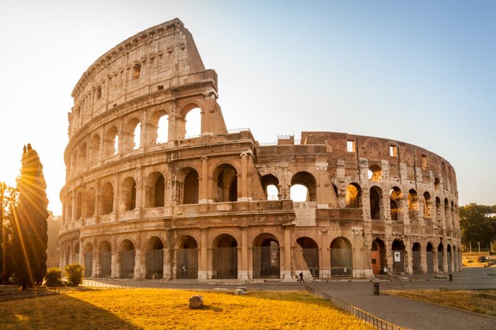 The Colosseum stands illuminated by sunset, surrounded by trees and a grassy area, casting long shadows on the ground.