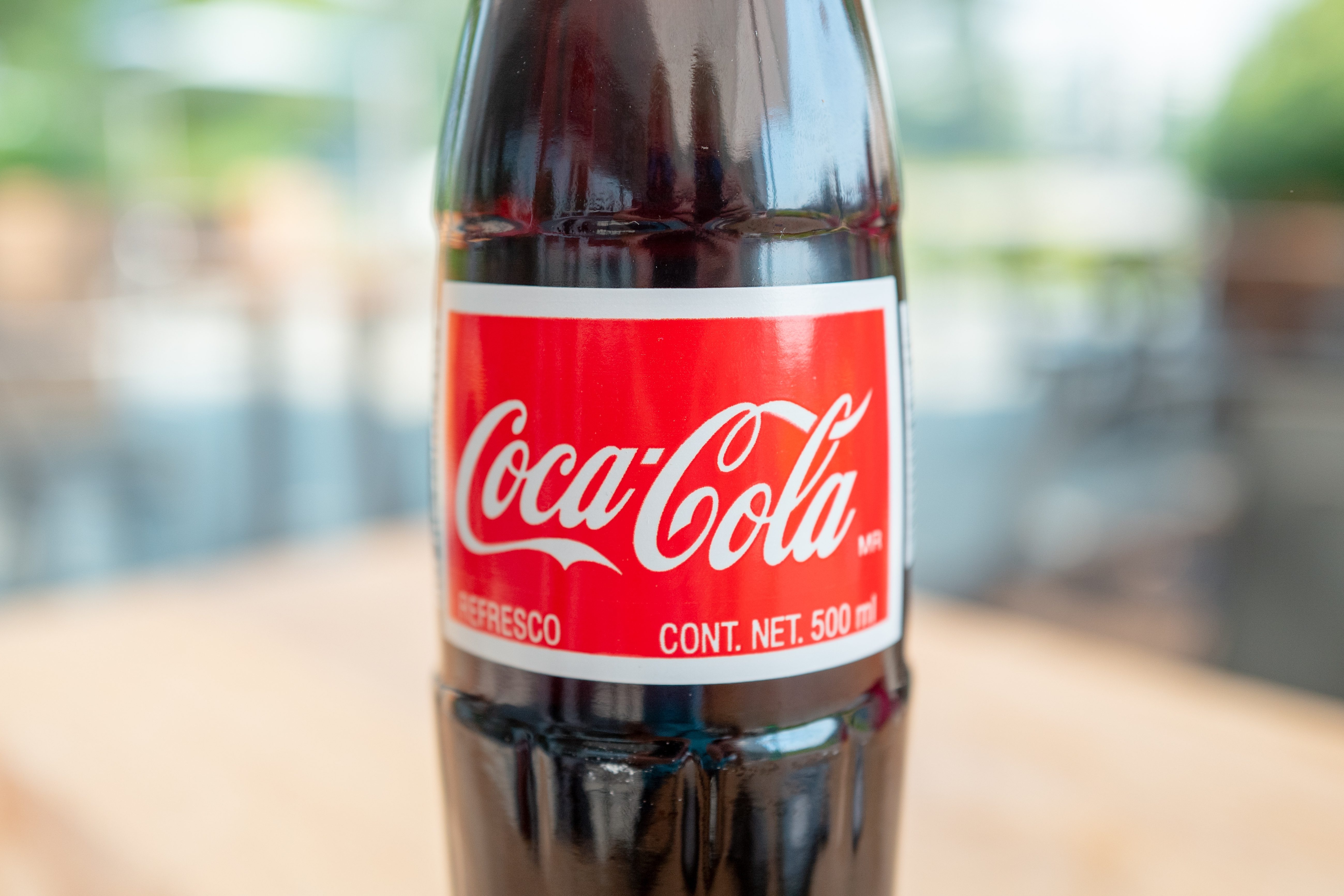 A Coca-Cola bottle stands upright on a blurred, outdoor table. Text: "Coca-Cola. Refresco. Cont. Net. 500 ml."