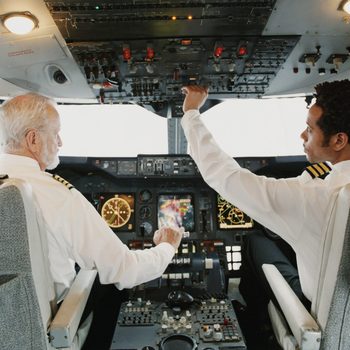 Pilots operate controls in a plane cockpit, surrounded by instruments, gauges, and switches.