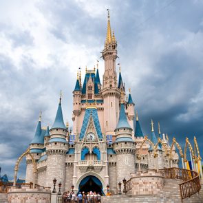 A large, ornate castle with blue spires, surrounded by people, stands beneath a dramatic, cloudy sky.