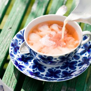 Milk being poured into a tea cup, creating swirls. The cup, adorned with blue floral patterns, rests on a green, weathered wooden table.