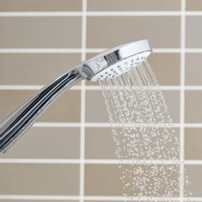 Shower head spraying water diagonally; background features beige tiled wall with brown and white grid pattern.