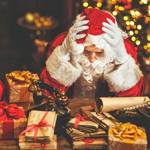 Santa sits stressed at a cluttered desk surrounded by gifts, letters, and vintage items in a festive holiday setting.