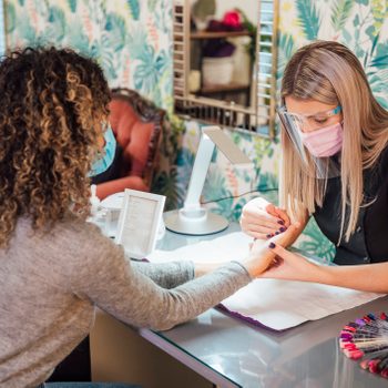 Manicurist filing customer's nails in a salon, surrounded by vibrant leaf-patterned wallpaper. A display with nail polish color samples is on the table.