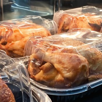 Rotisserie chickens sit packaged in clear plastic containers, displayed under warm lighting in a grocery store setting.