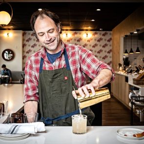 A person in a plaid shirt pours a drink from a shaker at a well-lit, modern restaurant bar.