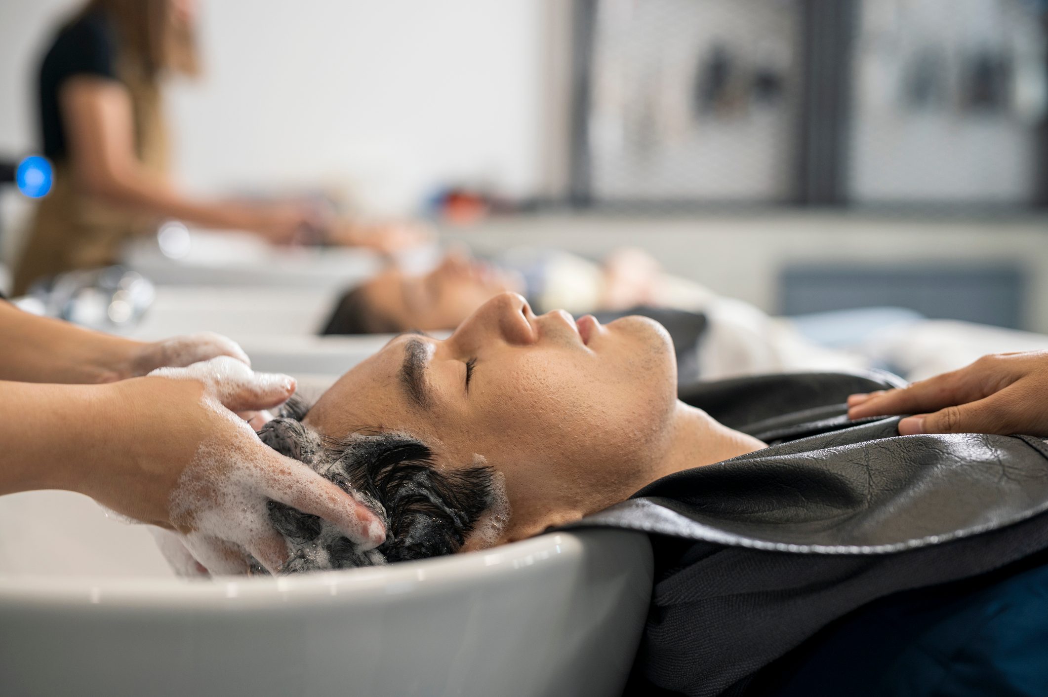 Person reclines, receiving a hair wash from someone else, surrounded by a salon environment with other clients and stylists in the background.
