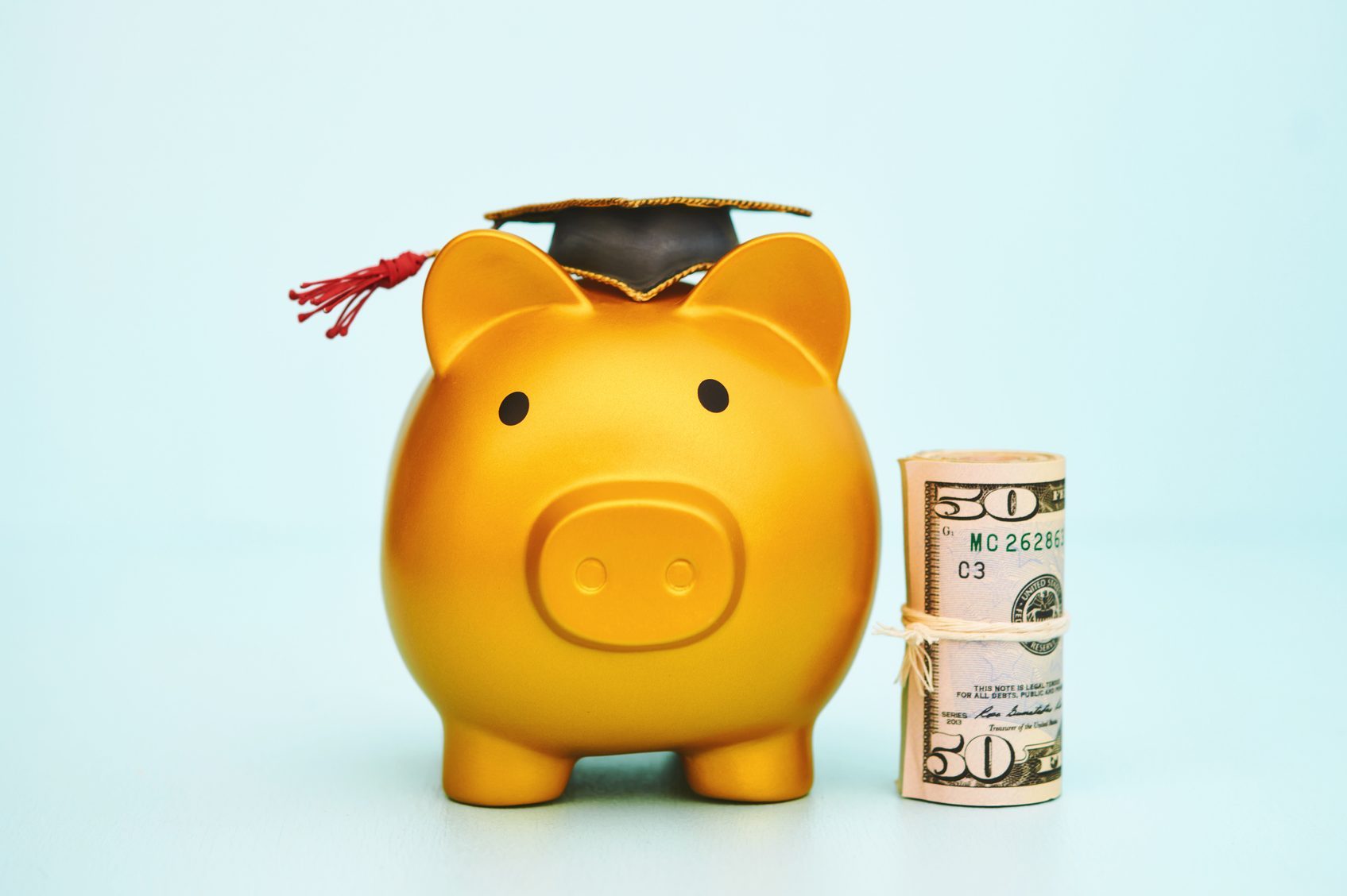 Golden piggy bank wears graduation cap, sitting next to a rolled stack of fifty-dollar bills, against a light blue background.