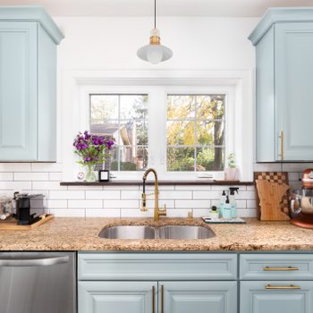Kitchen sink sits beneath large window; surrounded by granite countertops, pastel cabinets, and various kitchen tools. Bright, organized space with natural light.