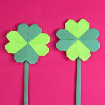Two origami clovers stand against a bright pink background.