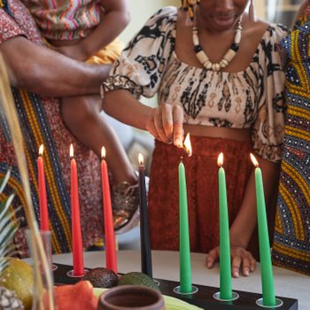 Candles being lit by a person, surrounded by colorful fabrics, a child held nearby, with fruits and decorations on a table below.
