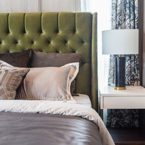 Bed displays plush pillows against an olive headboard, next to a nightstand with a lit lamp; draped curtains frame the elegant room.