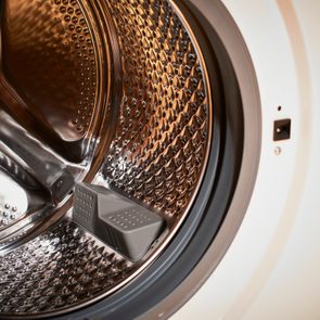 Washing machine drum, open, viewed from the front with shiny metallic surface and small perforations, situated in a white casing.