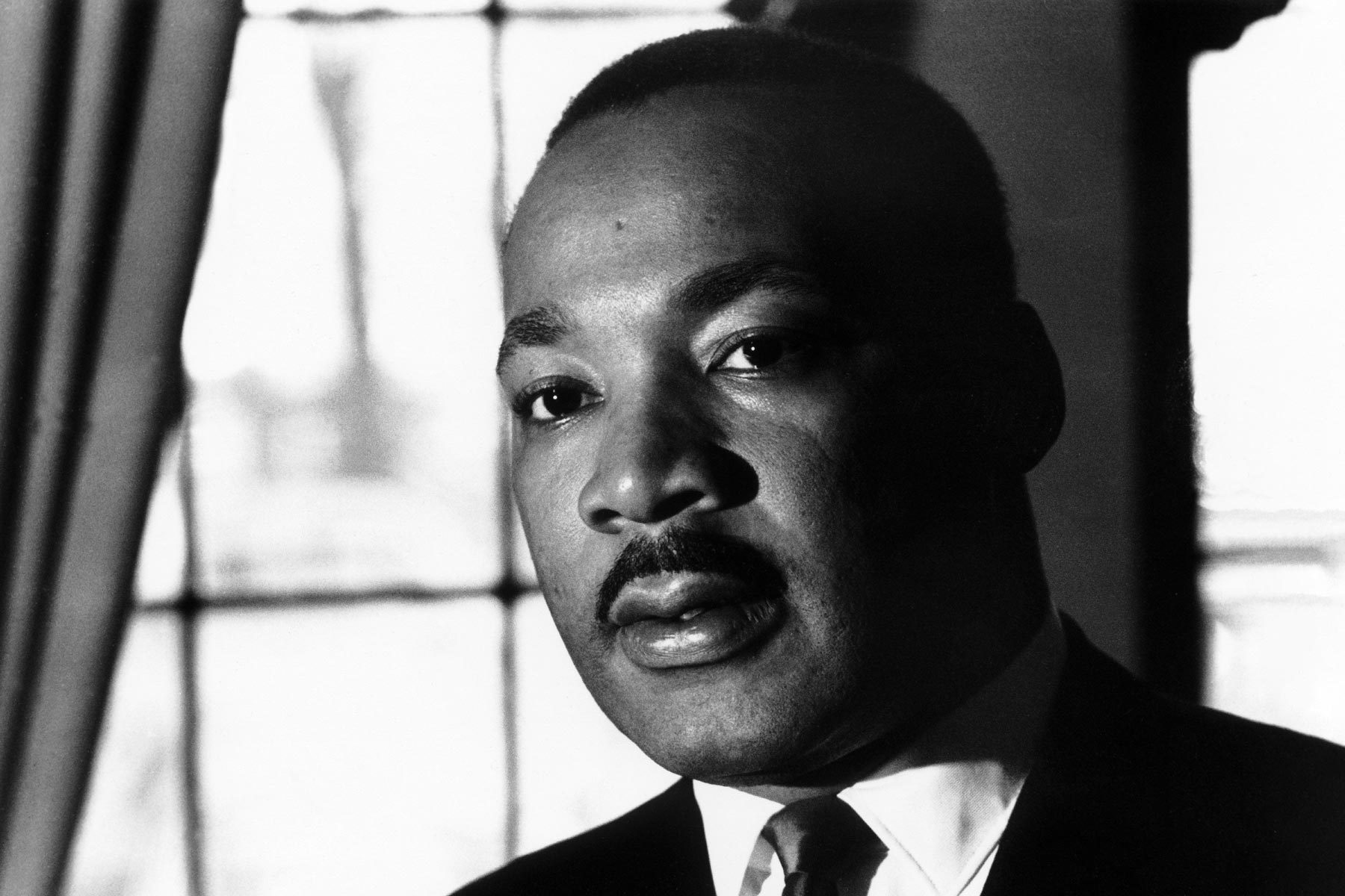 Person gazing solemnly, framed by a window with light filtering through, wearing a suit and tie, creating a contemplative and focused atmosphere.