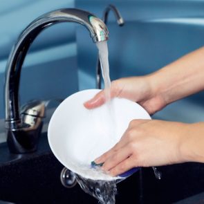 Hands are washing a white bowl under running water in a kitchen sink.
