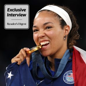 Athlete bites gold medal, showcasing pride, wearing a jacket with 