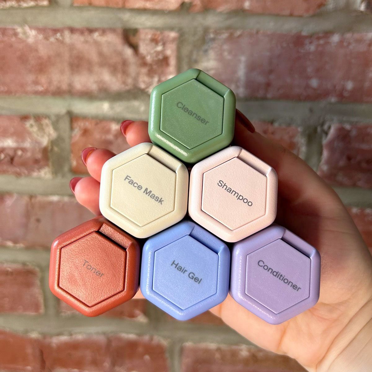 Hand holds six hexagonal containers labeled Cleanser, Face Mask, Shampoo, Toner, Hair Gel, and Conditioner; Reader's Digest Editor-Tested badge above. Brick wall background.