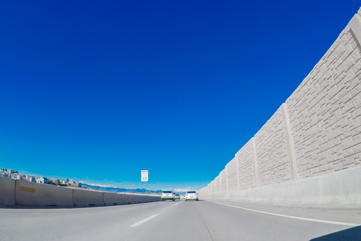 A wide highway under a clear blue sky, bordered by tall concrete sound barriers on both sides, with a few cars driving ahead.