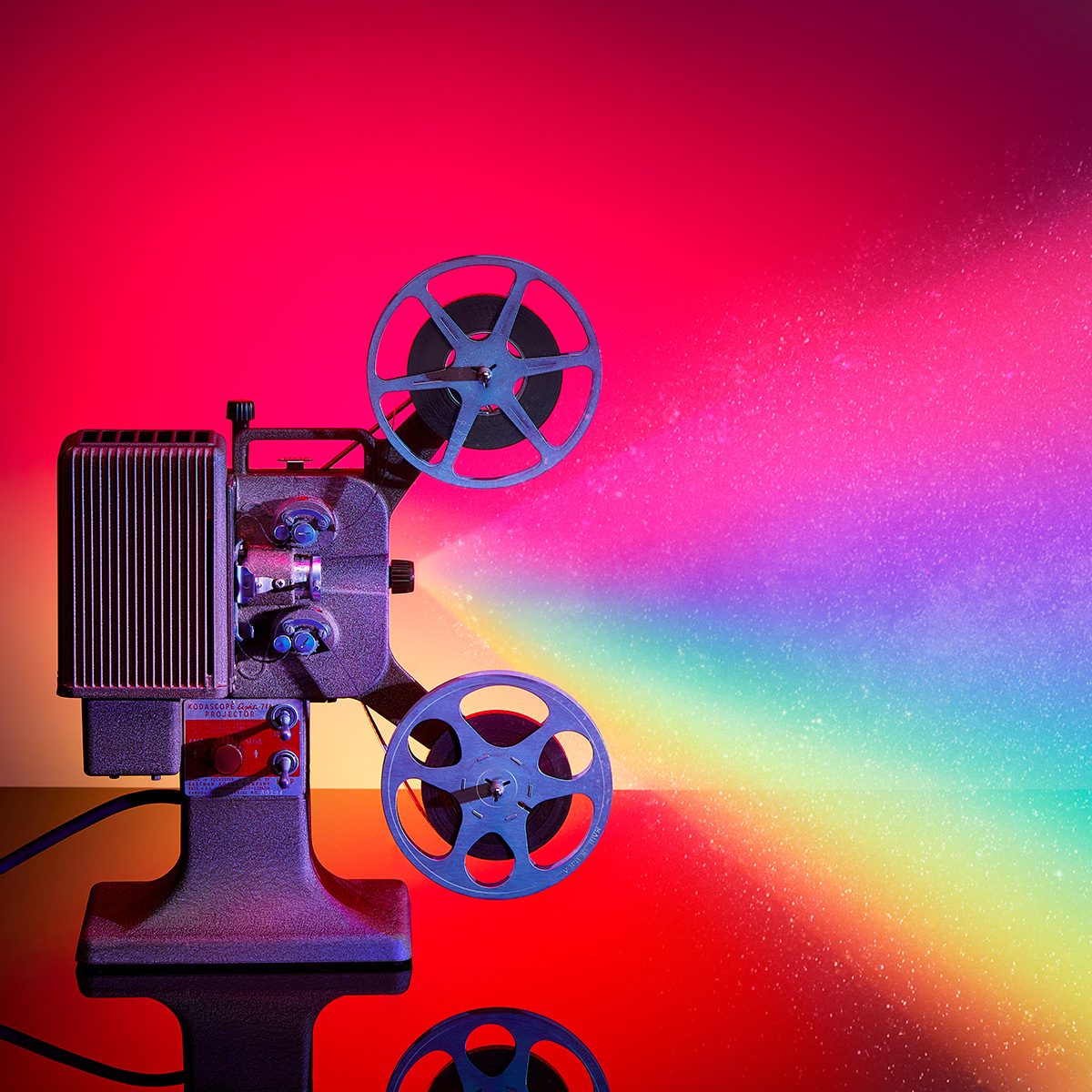 A vintage film projector displays a bright, colorful rainbow beam of light against a vibrant red and pink background.